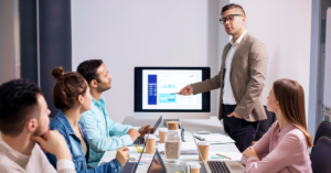 A group of employees sitting around a table listening to a data presentation being given.