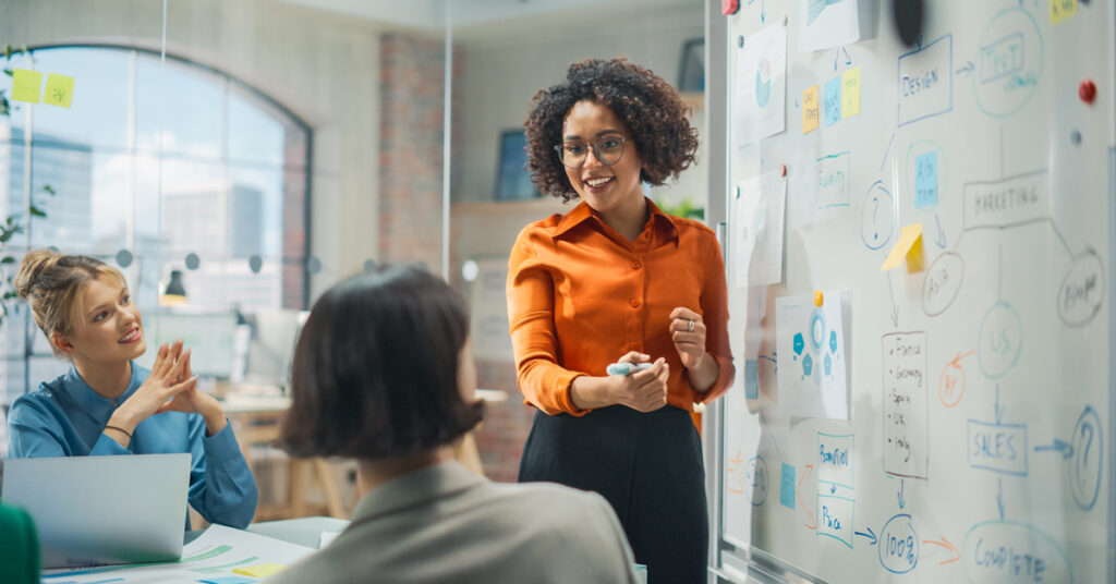 Woman wearing an orange shirt standing in front of a white board in conversation with colleagues